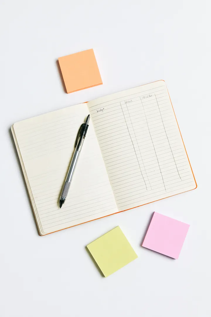 Open notebook with handwritten budget columns, a mechanical pencil and colored sticky notes on a white surface
